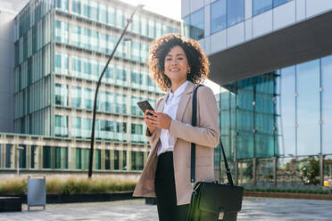 Beautiful hispanic businesswoman with elegant suit walking in the business centre- Adult female with business suit and holding mobile phone portrait outdoors - DMDF00602