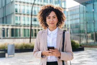 Beautiful hispanic businesswoman with elegant suit walking in the business centre- Adult female with business suit and holding mobile phone portrait outdoors - DMDF00604