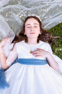 Top view of adorable teen girl in white dress and veil resting in green garden with closed eyes - ADSF46219