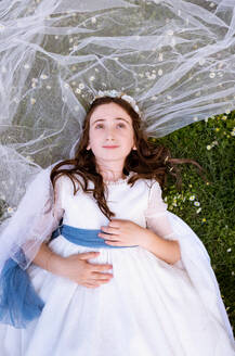 Top view of adorable teen girl in white dress and veil resting in green garden - ADSF46221