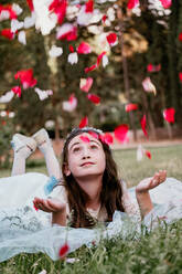 Preteen girl in white dress and headband lying on green lawn while looking up at falling flower petals of roses - ADSF46223