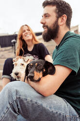 Side view of smiling bearded man embracing Dachshund dog while sitting on stairway with girlfriend and white Fox Terrier spending free time together - ADSF46225