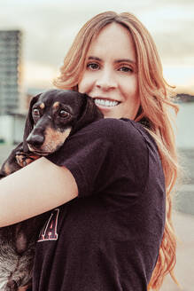 Young smiling redhead female owner with long hair hugging adorable Dachshund dog on city street against blurred background - ADSF46230