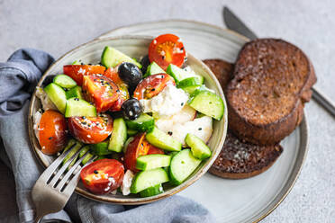 High angle of ceramic bowl with tasty healthy salad, vegetables and bread placed with sliced cucumber and tomato with cutlery and fabric over gray table - ADSF46240