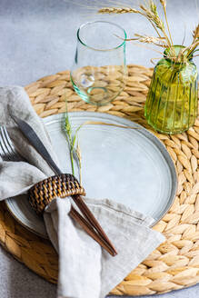 From above an elegant white ceramic plate with napkin-wrapped cutlery placed on a straw accent and a glass vase with ears of dried wheat - ADSF46246