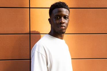 Portrait of serious African American male in white t-shirt looking at camera while standing against orange wall - ADSF46254