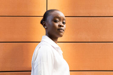 Confident young African American female in white shirt looking at camera while standing near orange wall - ADSF46257