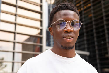 Serious African American male in trendy outfit and eyeglasses looking at camera while standing against modern buildings - ADSF46258