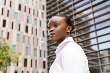 From below side view of young African American female in white shirt looking away while standing near modern buildings in city - ADSF46259