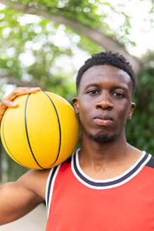 Portrait of confident young African American sportsman in activewear holding basketball on shoulder while looking at camera near blurred trees - ADSF46260