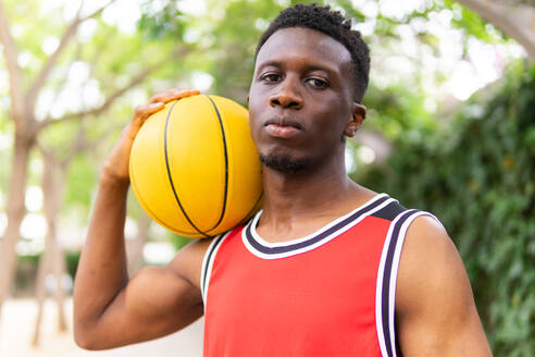 Portrait of confident young African American sportsman in activewear holding basketball on shoulder while looking at camera near blurred trees - ADSF46261