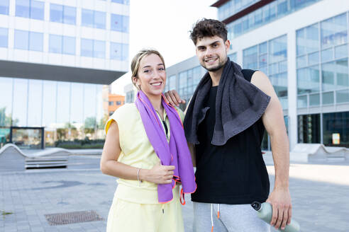 Smiling young couple with water bottle looking at camera while standing closely with hand on shoulder and towels around necks near blurred buildings - ADSF46263