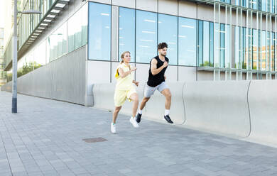 Focused athlete couple in sportswear and sneakers looking away while jogging on pavement near glass windows building during fitness exercise in city - ADSF46265