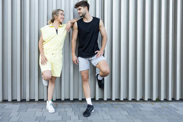 Full body of positive young couple in casual clothes smiling and looking at each other while standing and leaning on wall over paved street outdoors - ADSF46266