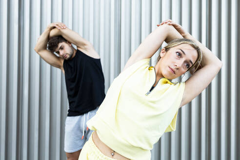 Serious young couple in casual clothes doing exercise while standing and swinging sideways on street by wall in daylight - ADSF46267