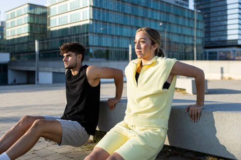Positive young couple in casual clothes doing sit down exercise on street with hands behind back on concrete bench while looking away by modern building - ADSF46268