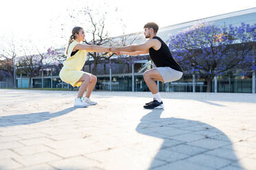 Full body side view of serious couple in sportswear and sneakers stretching arms and squatting while doing exercise workout on sports ground - ADSF46269