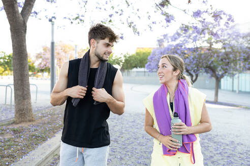 Smiling young couple with water bottle looking at each other while standing closely with and towels around necks near blurred park - ADSF46271