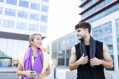 Smiling young couple with water bottle looking at each other while standing closely with and towels around necks near blurred buildings - ADSF46273