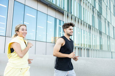 Focused happy athlete couple in sportswear while jogging on pavement near glass windows building during fitness exercise in city - ADSF46275