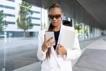 Confident young African American female in white formal suit standing near column of modern building with folder and phone looking at screen - ADSF46277