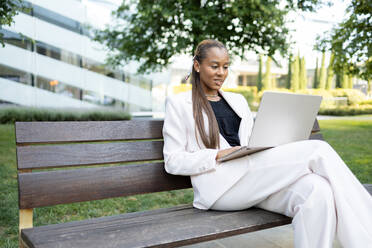 Smiling African American businesswoman with ponytail in white jacket and trousers sitting on wooden bench with crossed legs in city park and working on laptop - ADSF46279
