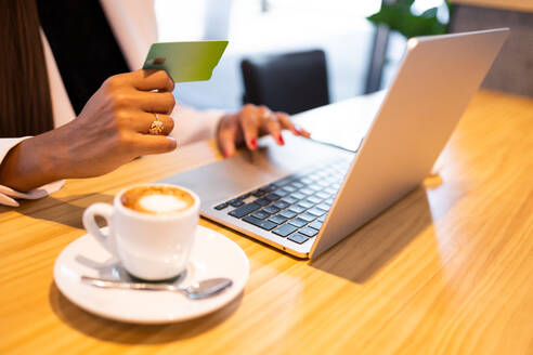 Anonymous young African American businesswoman in sitting at table near cup of coffee and entering credit card credentials in office - ADSF46281