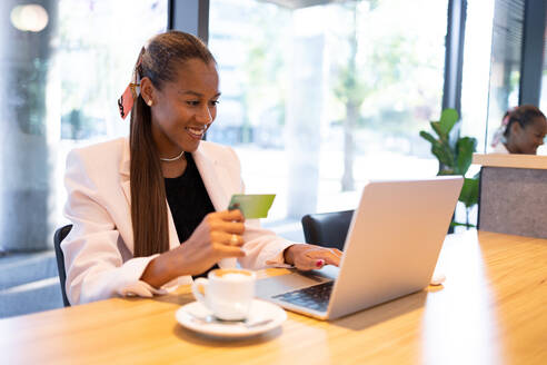 Smiling young African American businesswoman in white blazer sitting at table near cup of coffee and entering credit card credentials in office - ADSF46282