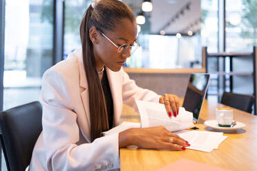 Side view of concentrated African American businesswoman in glasses and formal clothes sitting at table near laptop and cup of coffee while reading documents in office - ADSF46283