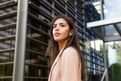 From below side view of pensive young Middle Eastern businesswoman in formal clothes standing near urban glass building and looking away - ADSF46288