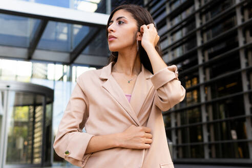 From below pensive young Middle Eastern businesswoman in formal clothes standing near urban glass building and looking away - ADSF46289