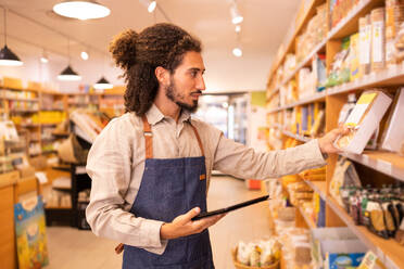 Side view of focused young ethnic male worker in casual clothes and apron standing near shelves while checking goods in modern grocery shop - ADSF46296