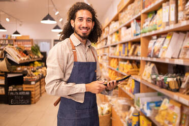 Portrait of happy ethnic male worker with long curly hair in casual clothes standing near shelves looking at camera smiling while working on tablet in modern lighted grocery shop - ADSF46298