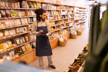 Portrait of serious ethnic male worker with long curly hair in casual clothes standing near shelves and working on tablet in modern lighted grocery shop - ADSF46300