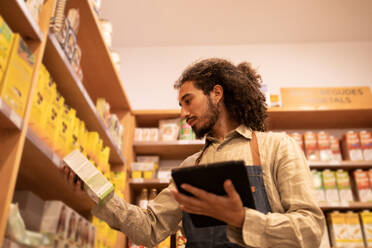 From below side view of focused young ethnic male worker in casual clothes and apron standing near shelves while checking goods in modern grocery shop - ADSF46302