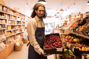 Side view of friendly ethnic male seller with curly hair smiling while showing box of fresh ripe plums standing at counter with assorted fruits and vegetables in supermarket - ADSF46303