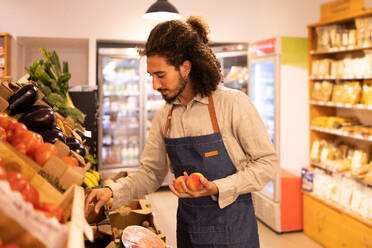 Positive young ethnic male seller with curly hair and beard in apron picking fresh ripe peaches while standing at counter with assorted fruits and vegetables in grocery store - ADSF46304