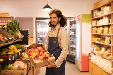 Side view of positive young bearded ethnic male seller with long curly hair in apron smiling and looking at camera while demonstrating box with fresh ripe peaches in supermarket - ADSF46305