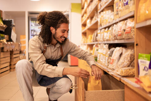 Side view of cheerful young bearded ethnic male worker with curly hair in apron sitting on haunches and smiling while choosing packs of food in supermarket - ADSF46308