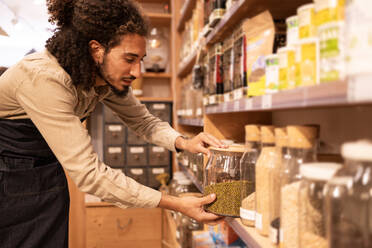 Side view of young ethnic bearded male seller with curly hair in denim apron standing in supermarket with glass jar of bulk food during work - ADSF46310