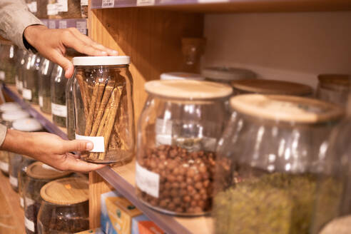 Crop unrecognizable male worker putting glass jar with cinnamon sticks on wooden shelf during work in grocery market - ADSF46313