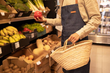 Cropped unrecognizable male seller in apron choosing fresh ripe bell pepper standing near assorted vegetables and fruits with straw basket in supermarket - ADSF46314