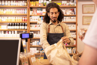 Positive young ethnic male seller with curly hair and beard in uniform smiling while putting products in eco friendly bag near crop client during work in store - ADSF46315