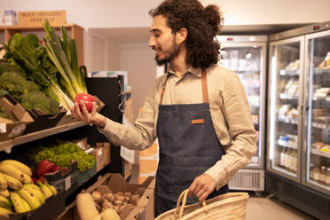 Positive young ethnic male seller in apron with curly hair and beard smiling while choosing fresh ripe bell pepper standing near assorted vegetables and fruits with straw basket in supermarket - ADSF46316