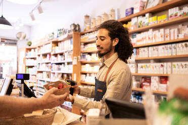 Side view of young bearded ethnic male seller with curly hair in apron scanning code of fresh ripe strawberries in eco friendly bowl during work in supermarket - ADSF46317