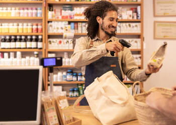 Cheerful young ethnic male cashier scanning barcode on pack of product and smiling near crop anonymous customer standing at table with weaved straw basket while working in eco friendly store - ADSF46318