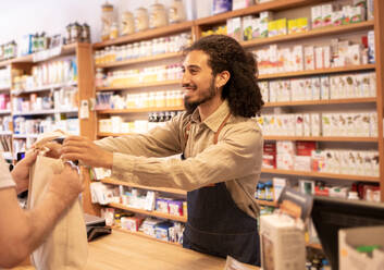 Cheerful bearded male in casual clothes with apron standing at table by drug shelves while dispensing drugs in bag to crop customer in pharmaceutical shop - ADSF46319