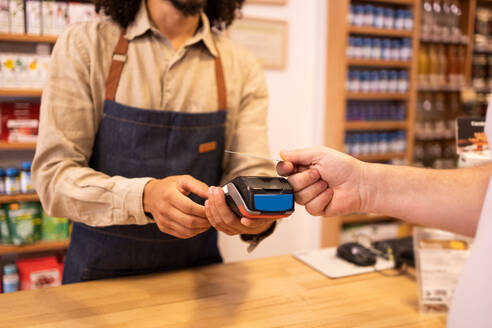 Crop anonymous male worker in casual cloth with apron receiving payment from client with POS machine and credit card while working in drugstore - ADSF46320