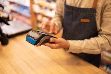 Crop anonymous male in casual clothes standing at wooden table while operating and processing financial payment on POS machine in shop - ADSF46322