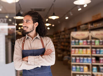 Portrait of positive bearded man in apron smiling and looking away while standing with arms crossed near dark blurred lighted background in supermarket - ADSF46327